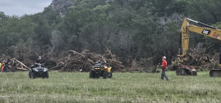 ATVs and an excavator clear debris along a tree line