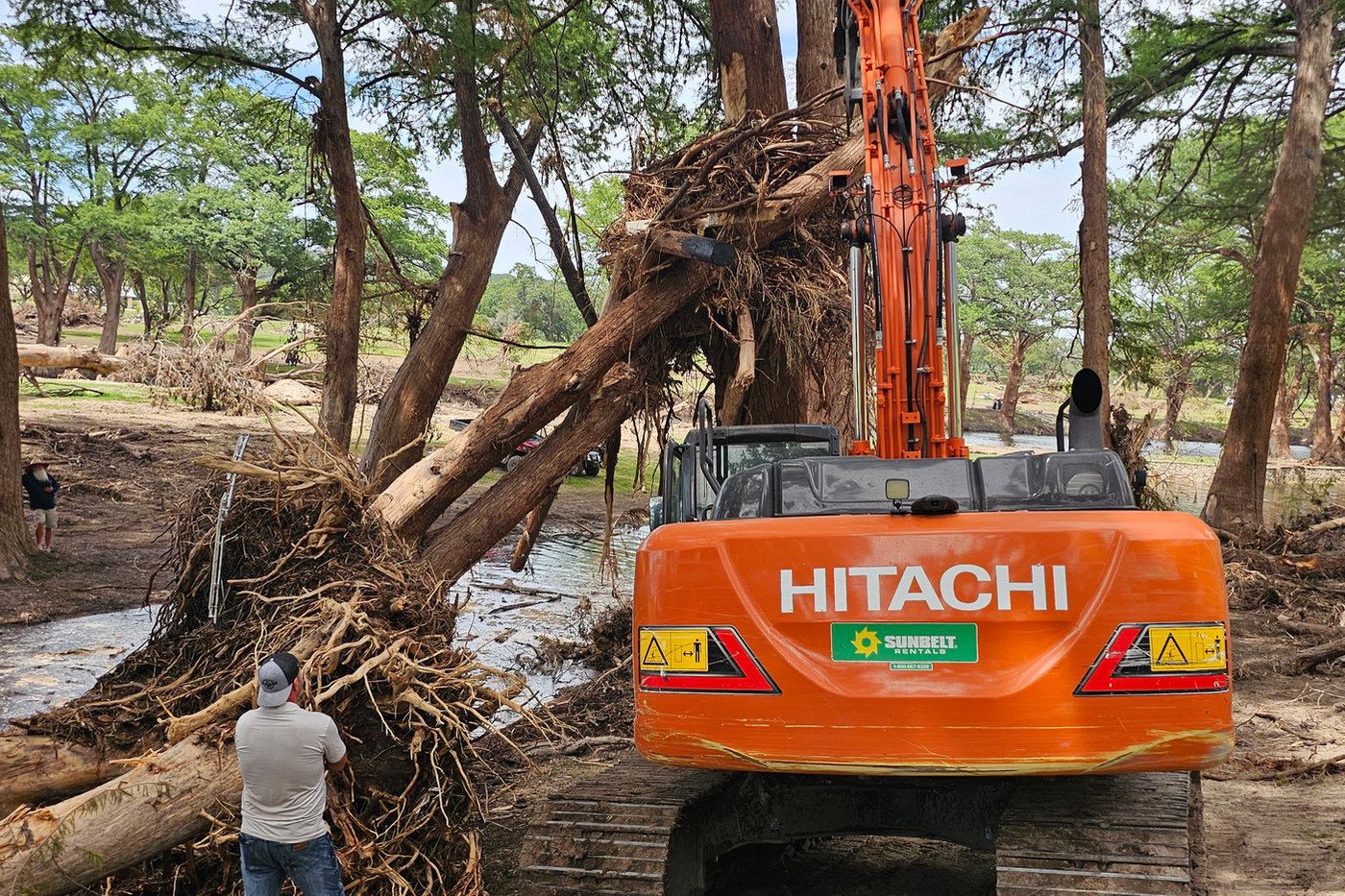 An excavator clears storm debris beside a river as volunteers look on